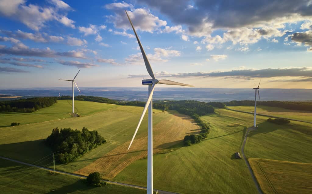 Aerial view of wind turbine farm. Wind power plants in green landscape against sunset sky with clouds. Aerial, drone inspection of wind turbine.